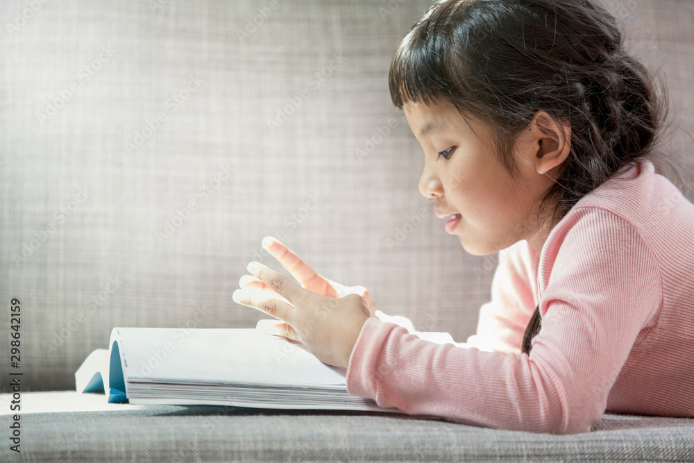 Cute little girl in casual clothes is counting numbers from lessons in the book on sofa in room. Cute little girl in casual clothes is counting numbers from lessons in the book on sofa in room.