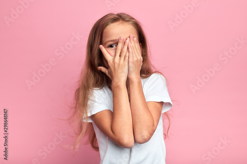 emotional little girl covers her face with her hand isolated over pink background, child watching horror film, movie, reaction, facial expression. isolated pink background © the faces
