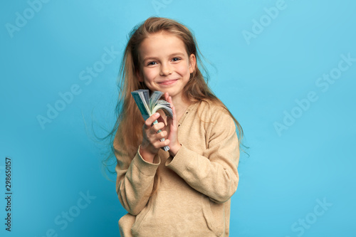 Cute cheerful happy little girl holding paper money, saving it, going to buy a phone isolated over blue, close up portrait, interest, happiness