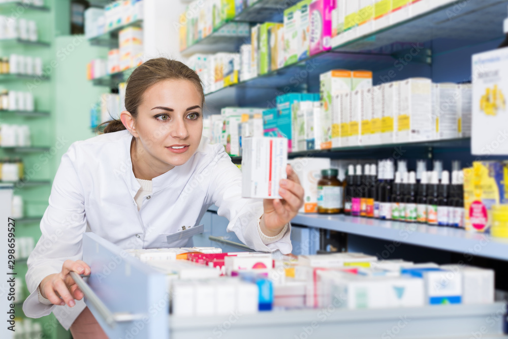 Young woman pharmacist is attentively looking medicines in apothecary