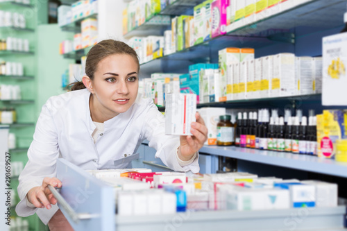 Young woman pharmacist is attentively looking medicines in apothecary