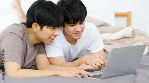 Happy young asian gay man couple using laptop computer while lying on bed at home, homosexual and lgbt with technology lifestyle concept