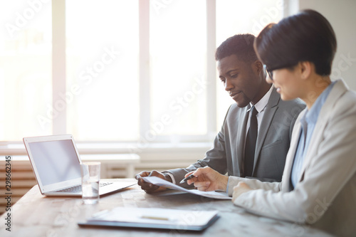 Side view portrait of young African businessman reading documents with female colleague while sitting at workplace against window in office, copy space