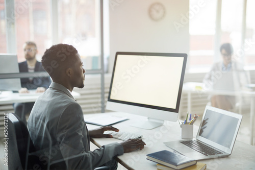 Side view portrait of African-American office worker using computer with black screen while sitting at workplace, shot from behind glass, copy space