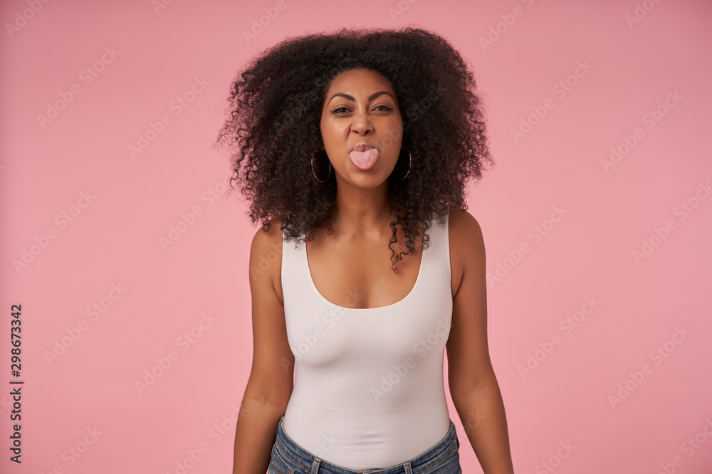Portrait of young dark skinned female with casual hairstyle wearing white shirt and jeans, looking at camera jofully and showing tongue, isolated over pink background Portrait of young dark skinned female with casual hairstyle wearing white shirt and jeans, looking at camera jofully and showing tongue, isolated over pink background