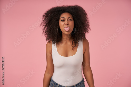 Portrait of young dark skinned female with casual hairstyle wearing white shirt and jeans, looking at camera jofully and showing tongue, isolated over pink background