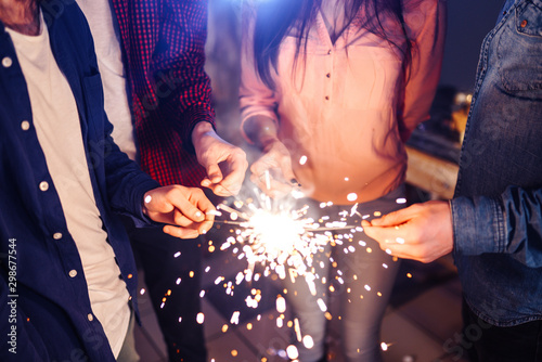 Group of happy people holding sparklers at party and smiling. Young people celebrating New Year together. Friends lit sparklers. Friends enjoying with sparklers in evening. Blur Background.
