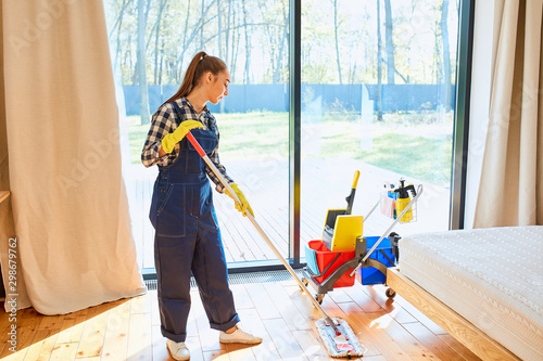 Enjoying cleaning service job. Young beautiful janitor cleaner in blue uniform with cleaning equipment stand mopping the floor in bedroom. Big panoramic window background