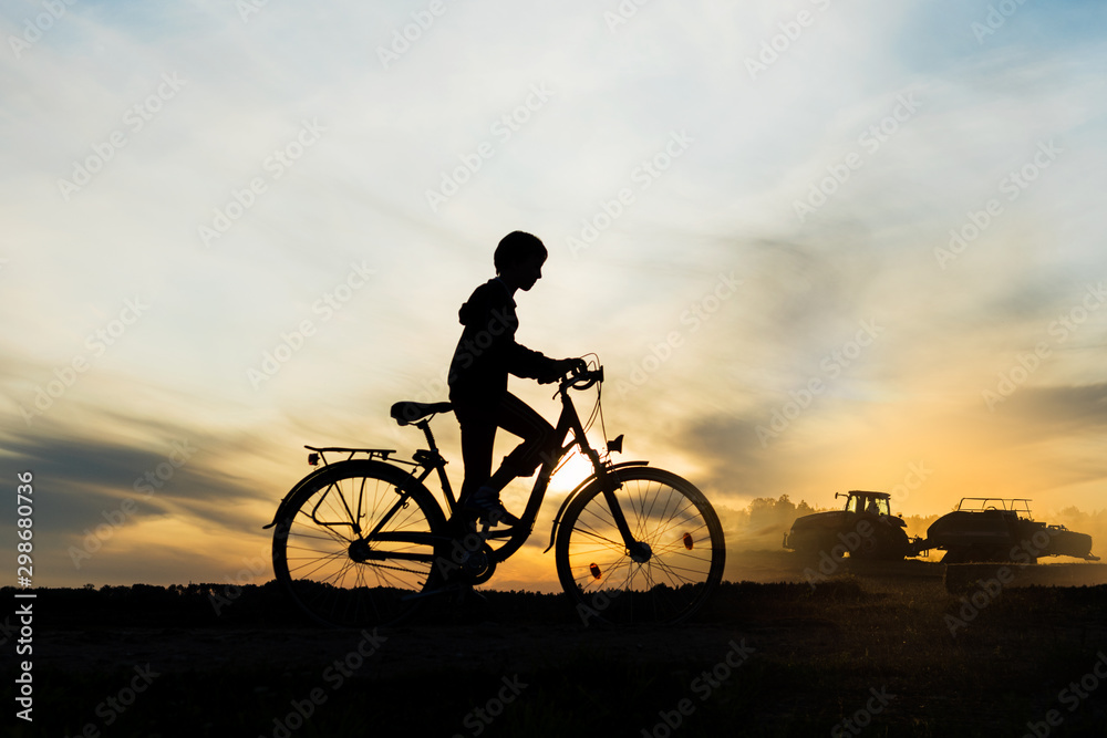 Obraz premium Boy , kid 10 years old riding bike in countryside, tractor working in background, silhouette of riding person and machine at sunset in nature
