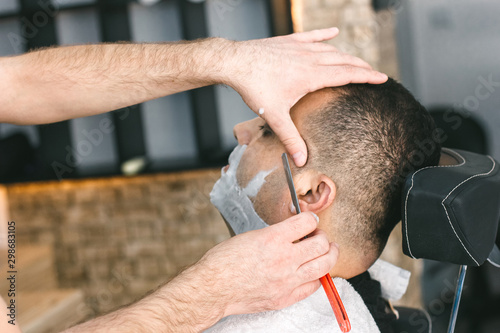 Hairdresser shaves a beard to a man with a razor in the barbershop