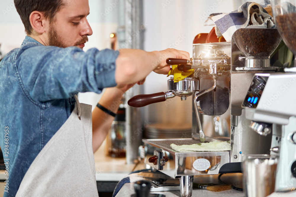 Confident self assured young barista, wears blue jeans shirt and white nest apron, press aromatic fresh coffee, uses tamper, cleans with rag, looks away with serious calm, professional concept Confident self assured young barista, wears blue jeans shirt and white nest apron, press aromatic fresh coffee, uses tamper, cleans with rag, looks away with serious calm, professional concept