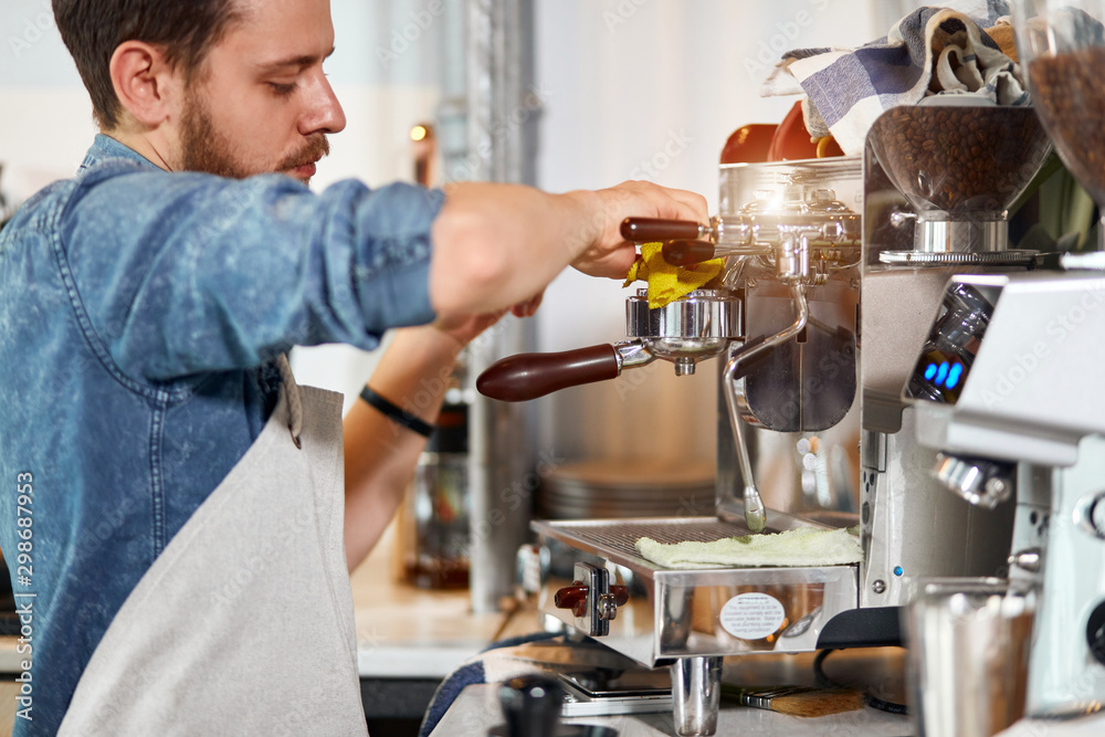 Confident self assured young barista, wears blue jeans shirt and white nest apron, press aromatic fresh coffee, uses tamper, cleans with rag, looks away with serious calm, professional concept