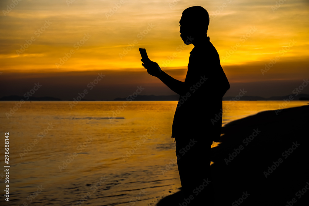 Silhouette of a man standing looking on a mobile phone by the sea after sunset.