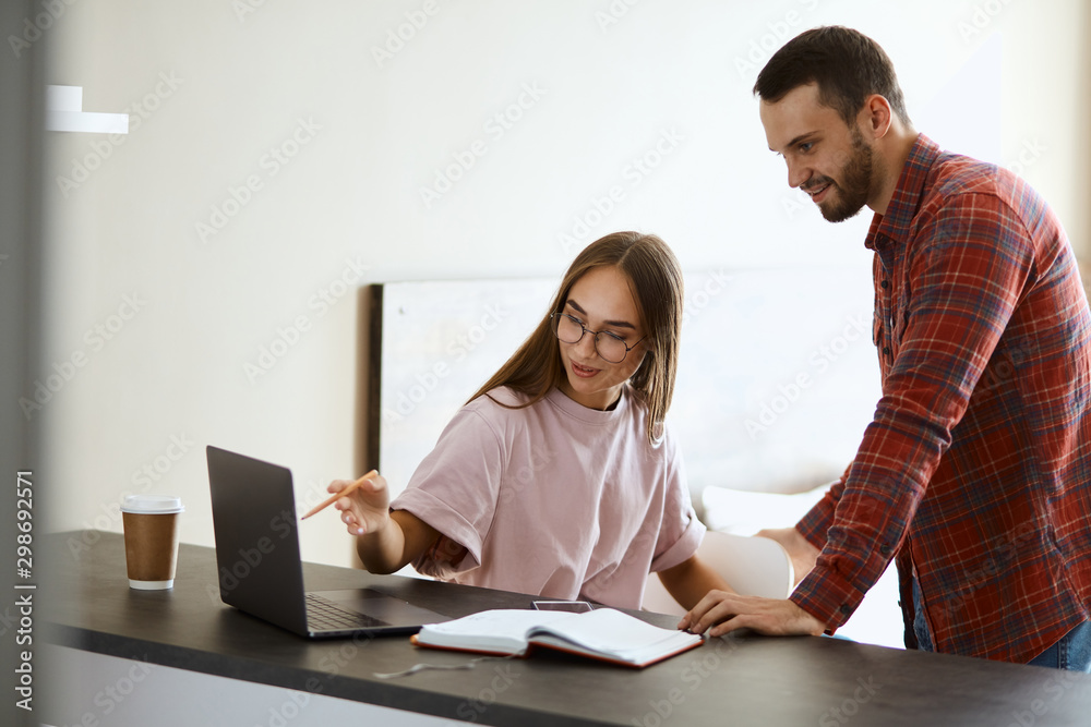 charming playful girlfriend points at screen of laptop computer, showing funny photo, sitting at black wooden table, friendship concept