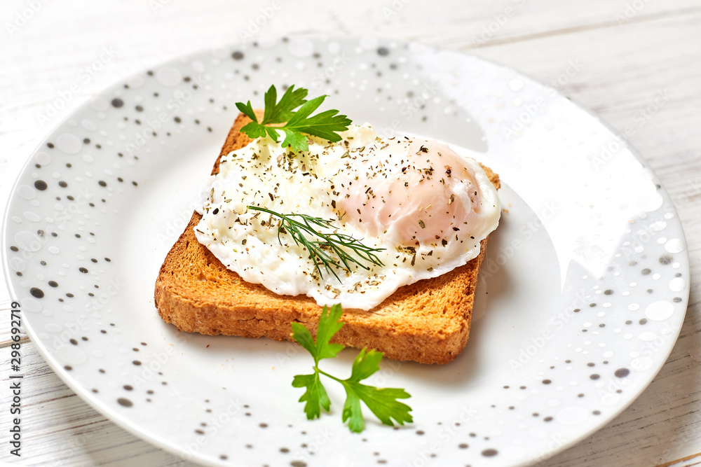 Poached egg, benedict on toasted white bread toast with salad and spices on a plate on a light wooden background.