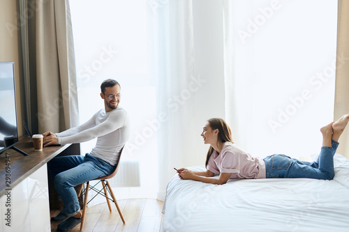Happy couple with laptop spending time together at home, pleasant man sits on chair, turns head at young wife, looks away positively, talking to pretty girl with pleasure, freelance concept, indoor