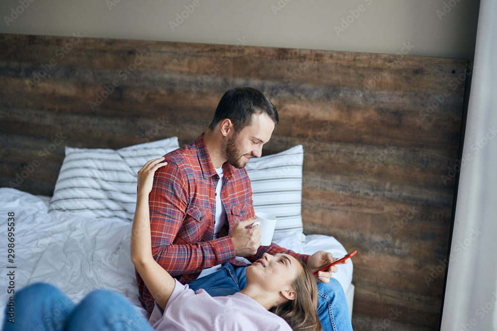Fototapeta premium romantic couple dressed in casual outfit, resting in bedroom, pretty girl lying on bed, putting head on knees husband, looking up with happy expression, stretches hand ut to hug delighted man