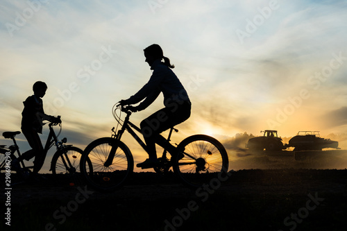 Boy , kid 10 years old, and girl riding bikes in countryside, tractor working in background, silhouette of riding persons and machine at sunset in nature