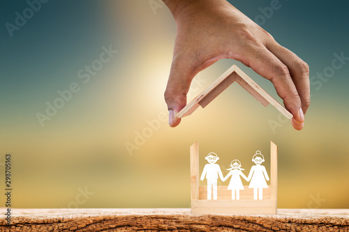 Mah hand hold to open a roof and wooden home with happy family of paper art is placed inside on nature sunlight, The saving money for house or business real estate owner in the future concept.
