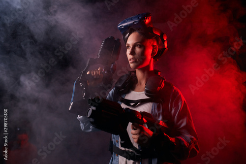 Attractive caucasian girl wearing special equipment, holding weapon for virtual game. Stand posing isolated over black background, red smoky neon backlit
