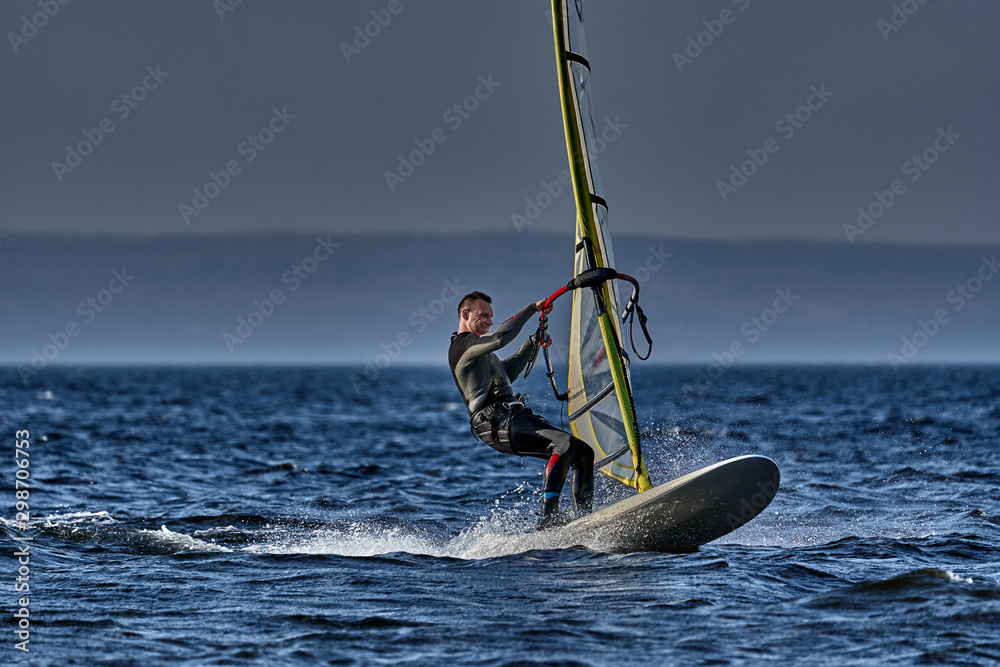 Naklejka premium A male athlete is interested in windsurfing. He moves on a Sailboard on a large lake on an autumn day.