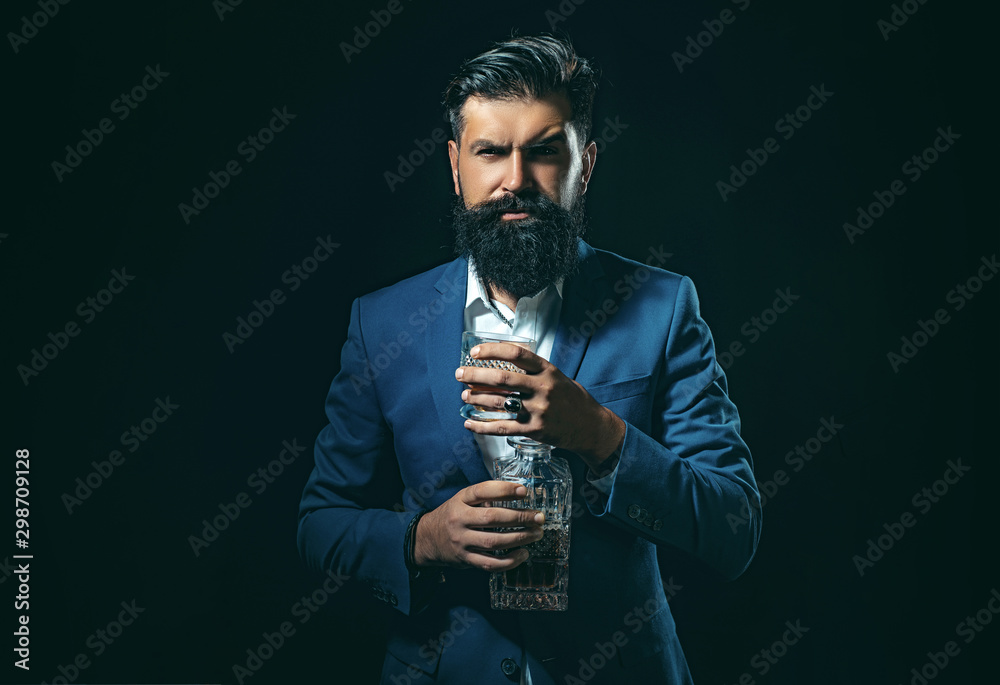 Naklejka premium Cheerful bearded man is drinking expensive whisky. Degustation of elite alcohol. Elegant and stylish man in classical wear holding glass with wiskey in hand.