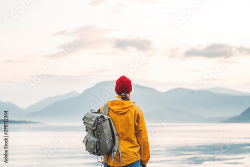 Back view of male tourist with rucksack standing on coast in front of great mountain while journey. Man traveler wearing yellow jacket with backpack explore scandinavia nature. Wanderlust outdoor
