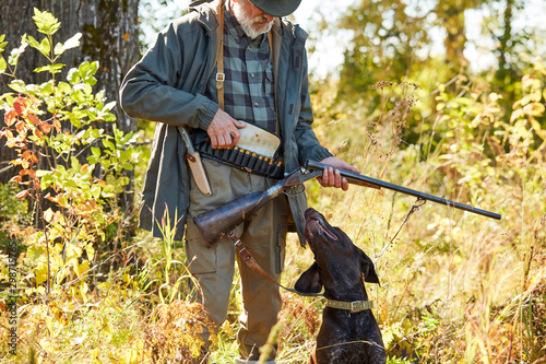 Black helper labrador with his owner hunter man in forest. Male loading shotgun