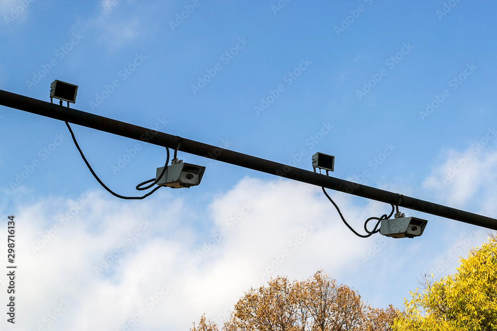 Two pairs of outdoor surveillance camera with sensor installed on a pole above a roadway. Modern automatic traffic control. Against a blue sky and yellow autunn trees. Two pairs of outdoor surveillance camera with sensor installed on a pole above a roadway. Modern automatic traffic control. Against a blue sky and yellow autunn trees.