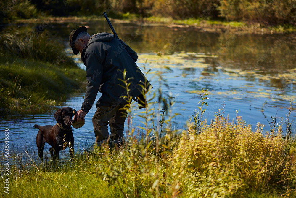 Hunter male with dog came to lake fot duck hunting. Autumn. Man give water to dog Hunter male with dog came to lake fot duck hunting. Autumn. Man give water to dog