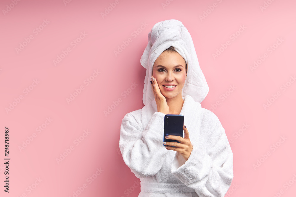 Smiling girl look at camera and chatting with friend dressed in bathrobe and in white towel after bathing. Isolated pink background. Health care, body care Smiling girl look at camera and chatting with friend dressed in bathrobe and in white towel after bathing. Isolated pink background. Health care, body care