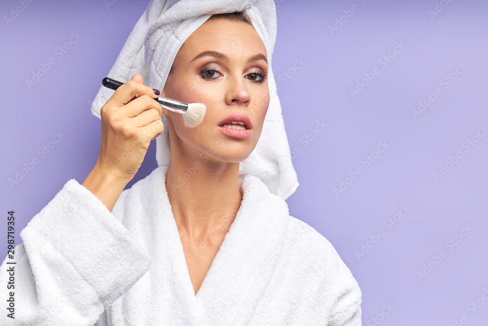 Young woman holding make up brushes, applying on face. isolated. Female in bathrobe and with towel on head. Beauty procedures concept Young woman holding make up brushes, applying on face. isolated. Female in bathrobe and with towel on head. Beauty procedures concept