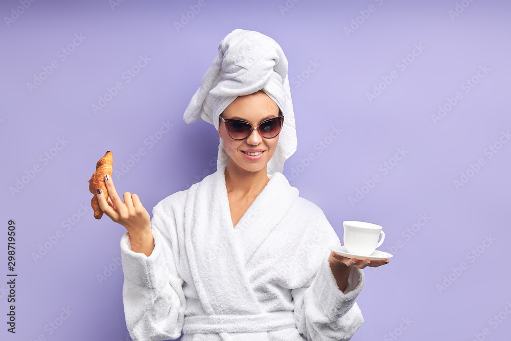 Close-up portrait of beautiful woman with cup of tea or coffee. Female dressed in bathrobe and towel on head after shower, sun glasses on eyes. Isolated violet background Close-up portrait of beautiful woman with cup of tea or coffee. Female dressed in bathrobe and towel on head after shower, sun glasses on eyes. Isolated violet background