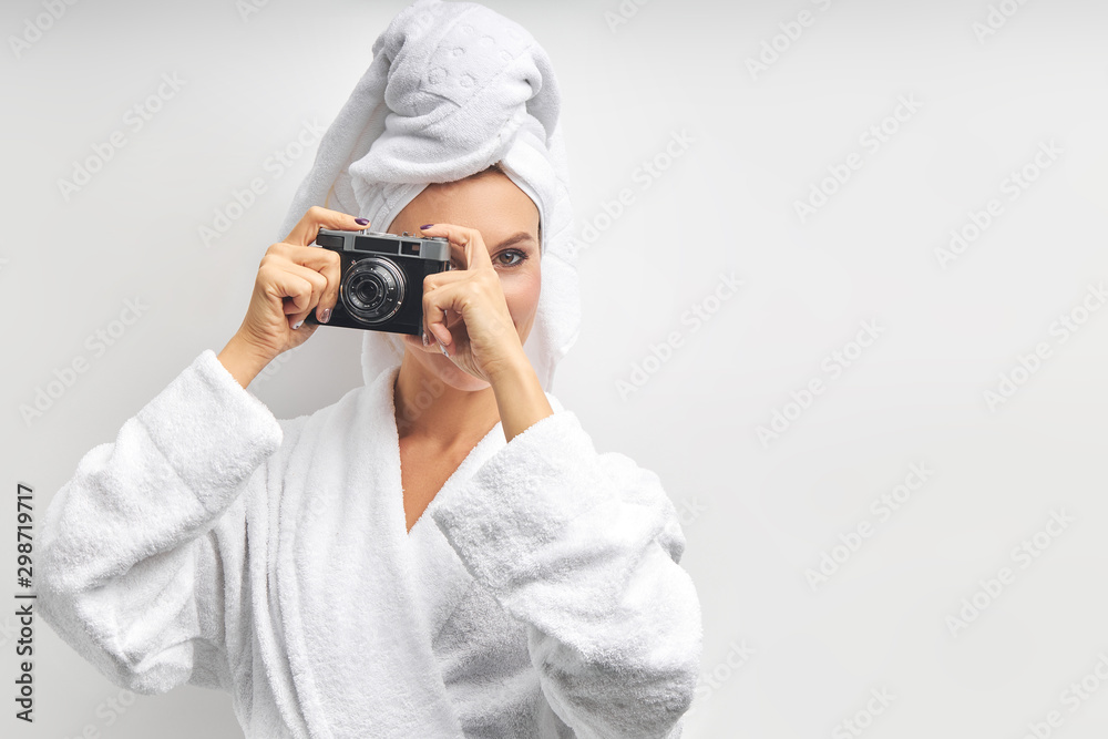 Caucasian pretty woman stand taking photo with photo camera, dressed in white bathrobe and towel on head, look at camera with one eye. White isolated background Caucasian pretty woman stand taking photo with photo camera, dressed in white bathrobe and towel on head, look at camera with one eye. White isolated background
