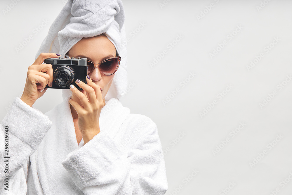 Caucasian pretty woman stand taking photo with photo camera, dressed in white bathrobe and towel on head. White isolated background