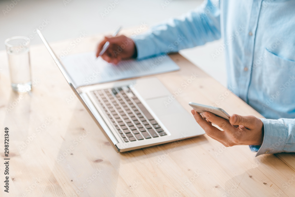 Young businessman in blue shirt using smartphone and laptop and making notes Young businessman in blue shirt using smartphone and laptop and making notes