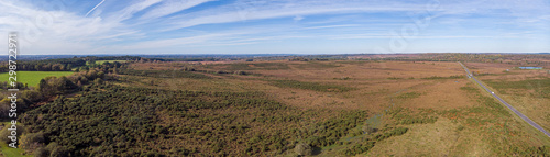 An aerial pamoramic view of the New Forest along a rural road with heartland, forest, pond and wild vegetation with beautiful autumn colors under a majestic blue sky and white clouds