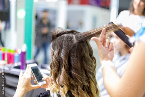 close up stylist makes a girl client styling hair waving with curling irons in a hairdresser. customer girl looks into smartphone in blur. close up, soft focus