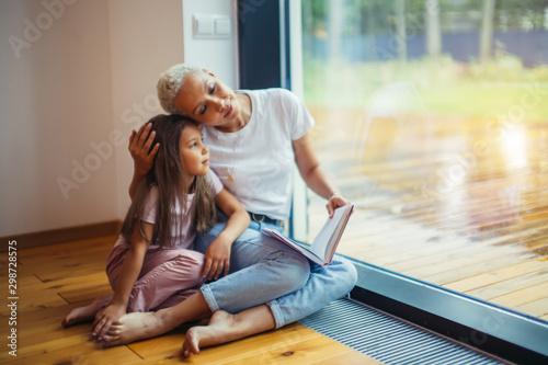Beautiful mother and her daughter sit on floor reading book near big panoramic window. White colors