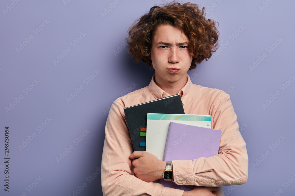 Tired bothered long haired boy with freckles, looks at camera with offended face, purses lips, knits impressive brows, dissatisfied by studying process, isolated portrait Tired bothered long haired boy with freckles, looks at camera with offended face, purses lips, knits impressive brows, dissatisfied by studying process, isolated portrait