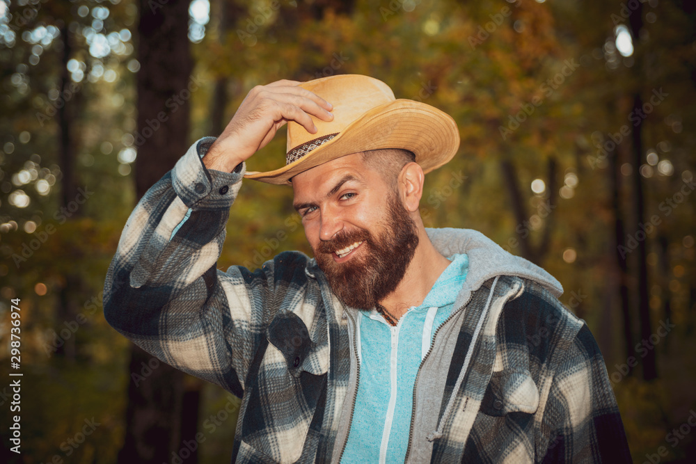 Smiling bearded macho man in cowboy straw hat and plaid hudie at autumnal forest background. An elegant man in greeting takes off his hat. Portrait of stylish fashion man. Smiling bearded macho man in cowboy straw hat and plaid hudie at autumnal forest background. An elegant man in greeting takes off his hat. Portrait of stylish fashion man.