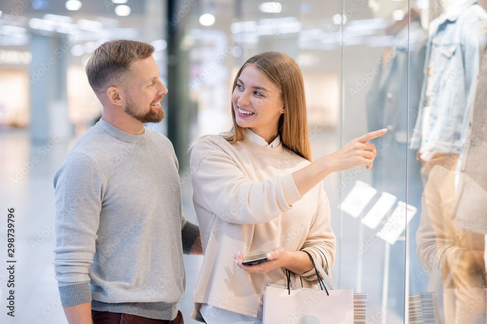 Young woman pointing at coat in shop window while looking at her husband Young woman pointing at coat in shop window while looking at her husband