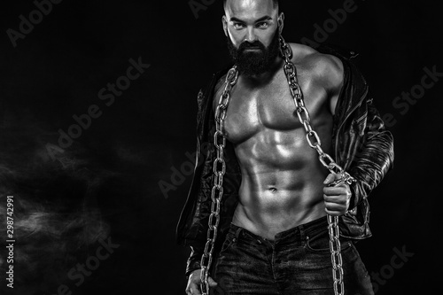 Men's fashion concept. Close-up portrait of a brutal bearded man topless in a leather jacket. Athlete bodybuilder on black background. © Mike Orlov