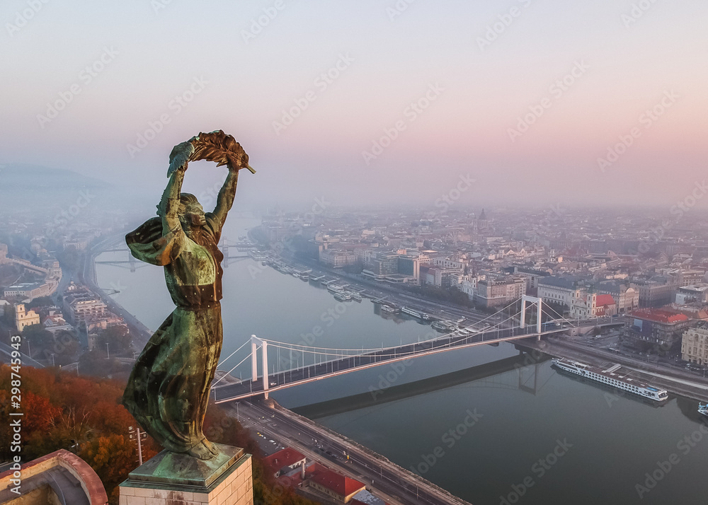 Naklejka premium Aerial view to the Statue of Liberty with Elisabeth Bridge and River Danube taken from Gellert Hill on sunrise in fog in Budapest, Hungary.