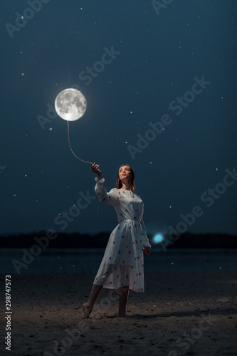 Young attractive girl in a long white dress holds the moon on a rope. Art photography