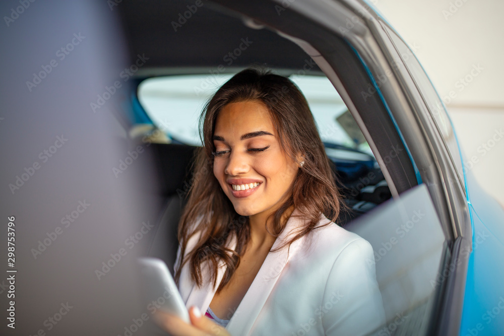 Young, beautiful woman sitting in the back seat of the car with a tablet in hand and drinking coffee. Close up portrait of a young business woman using digital tablet in the back seat of the car.