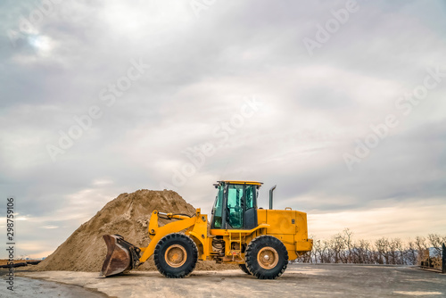 Bulldozer in front of a mound of soil with leafless trees and cloudy sky background