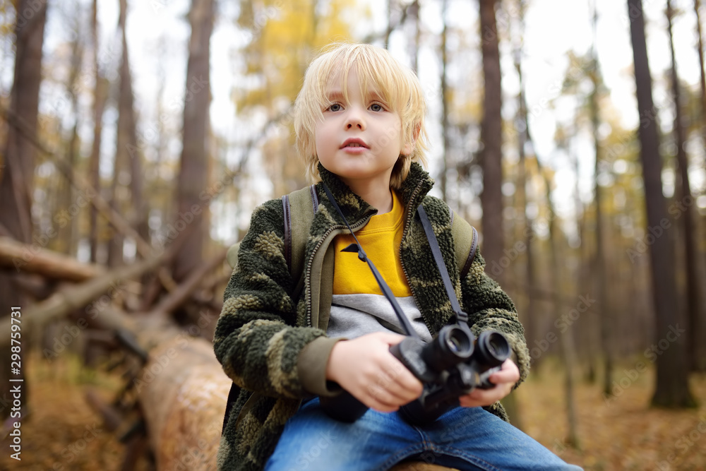 Little boy scout with binoculars during hiking in autumn forest. Child is sitting on large fallen tree and looking through a binoculars.