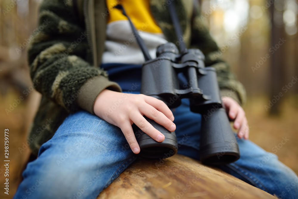Fototapeta premium Little boy scout with binoculars during hiking in autumn forest. Child is sitting on large fallen tree and looking through a binoculars.