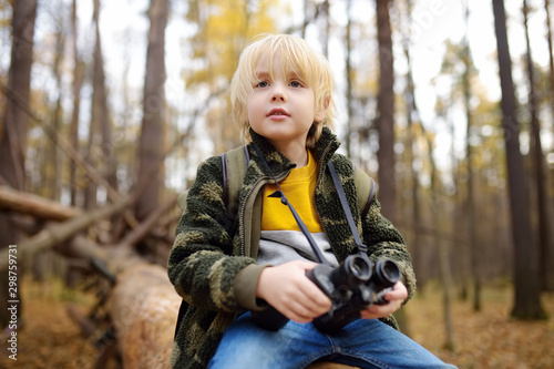 Little boy scout with binoculars during hiking in autumn forest. Child is sitting on large fallen tree and looking through a binoculars. © Maria Sbytova